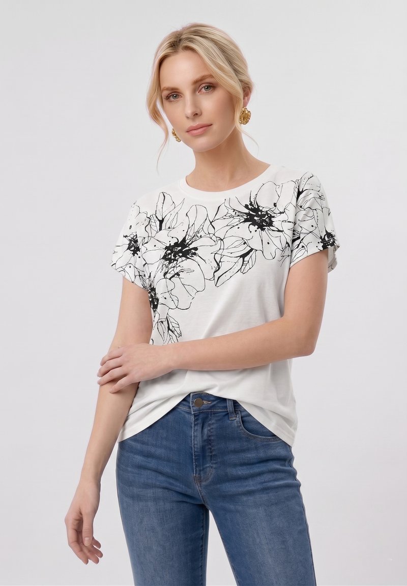 Femme blonde portant des boucles d'oreilles en or, un t-shirt blanc avec un imprimé floral noir et un jean bleu, posant bras croisés devant un fond clair.