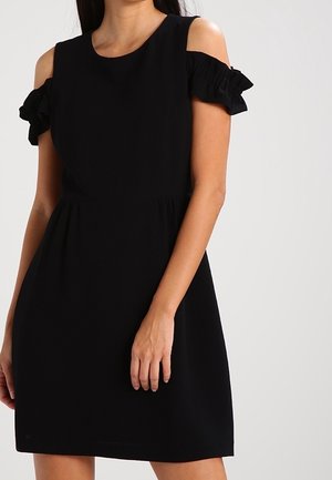 Kvinde iført en sort kold- skulder kjole med flæse-ærmer og en tætsiddende talje, stående foran en ensfarvet baggrund.