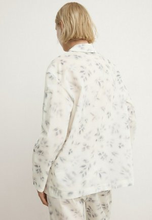 Persoană cu păr scurt și blond poartă o cămașă largă albă și pantaloni cu un model delicat de frunze gri, văzută din spate.