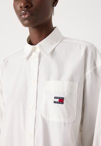 Hvit skjorte med spiss krage og venstre brystlomme som viser en rød, hvit og blå "TOMMY JEANS" -logo. Glatt tekstur.