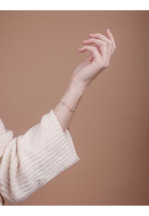 Jemný náramek z růžového zlata se třemi akcenty ve tvaru diamantu, nošený na štíhlém zápěstí, v kombinaci s krémově barevným pleteným svetrem.