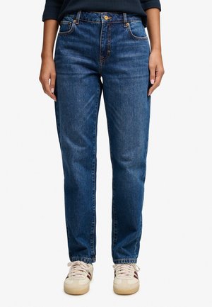 Persoon draagt blauwe straight-leg jeans en witte sneakers, staat met ontspannen handen langs het lichaam.