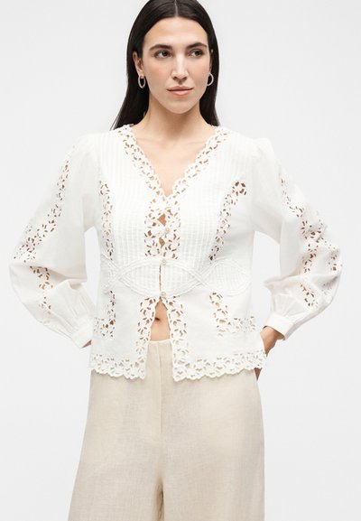 Γυναίκα με σκούρα μαλλιά φοράει λευκό μπλουζάκι με μακριά μανίκια, δαντελωτά κοψίματα και κουμπιά, σε συνδυασμό με ανοιχτό μπεζ παντελόνι με φαρδύ πόδι.