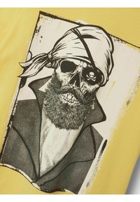 Camiseta de algodón amarilla con un gráfico negro de un cráneo con barba y bandana, situado dentro de un borde rectangular.