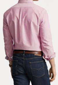 Jasnoróżowa koszula z długim rękawem i kołnierzykiem zapinanym na guziki, zestawiona z ciemnoniebieskimi dżinsami z kontrastującymi przeszyciami i tylnymi kieszeniami z akcentami wzorniczymi.