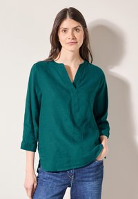 Türkisfarbene Leinenbluse mit V-Ausschnitt, drei Viertelärmeln und seitlichen Schlitzen. Kombiniert mit blauen Jeans. Glatte Textur und lässiges Design.