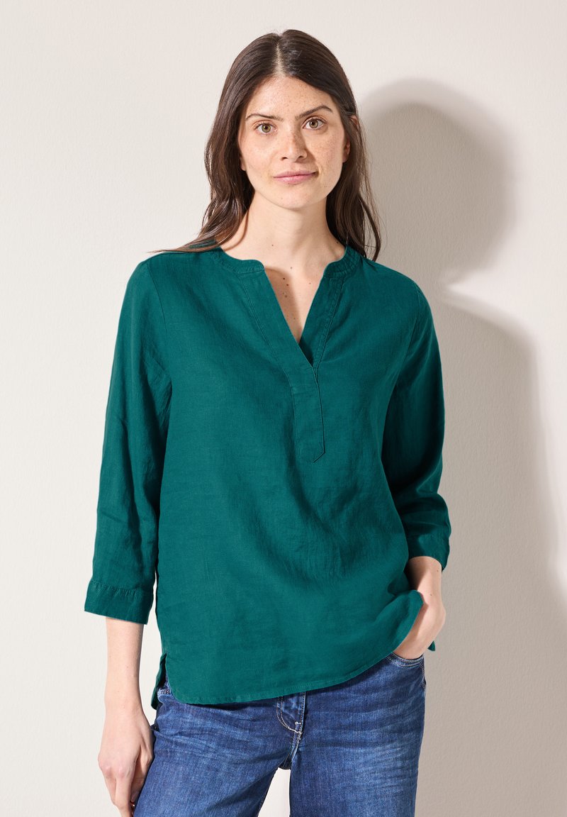 Türkisfarbene Leinenbluse mit V-Ausschnitt, drei Viertelärmeln und seitlichen Schlitzen. Kombiniert mit blauen Jeans. Glatte Textur und lässiges Design.