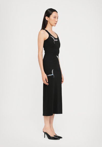 Rochie neagră fără mâneci, cu un corset ajustat și o lungime midi, prezentând motive brodate cu păsări. Asortată cu tocuri subțiri negre lucioase.