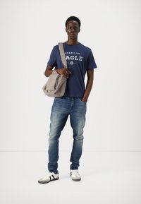 Temno modra majica s tiskom "American Eagle", svetlo siva torba, modri kavbojki in beli športni čevlji s črnimi črtami. Preprost sproščen videz.