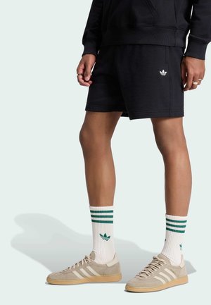 Osoba nosząca czarne spodenki Adidas, białe skarpetki Adidas z zielonymi paskami oraz beżowe buty sportowe Adidas stojąca na jasnym tle.