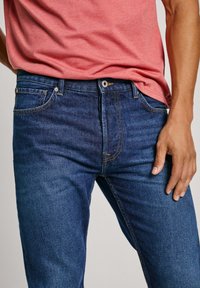 Jeans de mezclilla en azul oscuro con corte recto, con costuras decorativas, cremallera y bolsillos laterales. Llevados con una camiseta rosa.