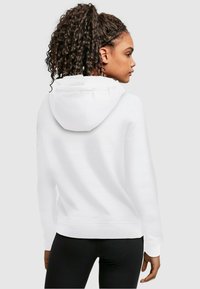 Fehér kapucnis pulóver laza szabással, bordás mandzsettákkal és hem-mel, puha anyagból készült, fekete leggings fölé húzva. Közepes méretű kapucnival rendelkezik.