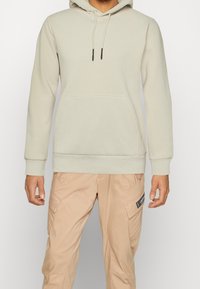 Felpa grigia chiara con tasca a canguro, cappuccio con coulisse e polsini a costine. Abbinata a pantaloni cargo beige con tasche applicate.