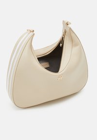 Beżowa skórzana torba na ramię o zaokrąglonym kształcie, złotymi akcentami, zamkiem błyskawicznym oraz białym paskiem na jednej stronie.