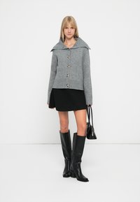 Cardigan gris en tricot avec un large col, des manches longues et une fermeture à boutons, assorti à une mini-jupe noire et des bottes noires jusqu'aux genoux.