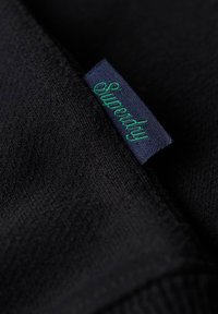 Tessuto nero con una consistenza morbida; un'etichetta di tessuto visibile riporta "Superdry" in verde su uno sfondo blu navy.