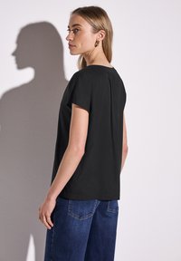 Svart kortärmad topp med rund halsringning och veckdetalj på baksidan, matchad med blå vida jeans. Slät textur och enkel design.