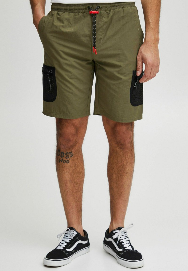 Khaki shorts med elastisk talje, justerbar snøre, to sidelommer og en struktureret sort lynlås-lomme. Hvide sneakers på.