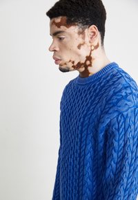 Fényes kék, kötött pulóver, textúrált mintákkal, bordás nyakkal és laza fazonnal.