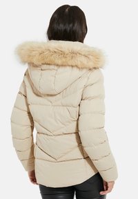 Beige Pufferjacke mit einer Kapuze aus Kunstfell, versehen mit gestepptem Muster und gerippten Bündchen. Das Design hat einen leicht abgerundeten Saum auf der Rückseite.