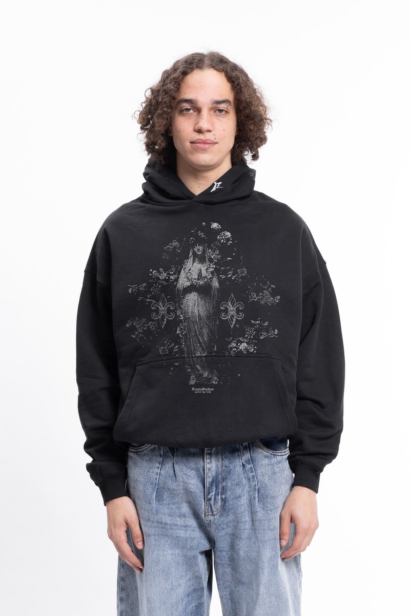Person with curly hair wearing black hoodie with religious statue graphic and light blue high-waisted jeans standing against white background.