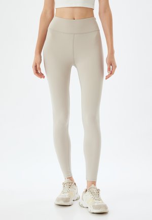 Lichtbeige high-waisted leggings met een gladde textuur en slim fit, gecombineerd met lichtgekleurde sportschoenen met gele accenten.