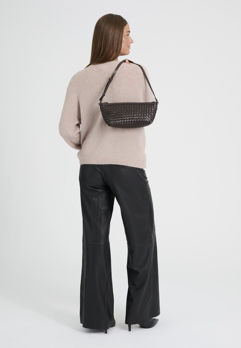 Femme portant un pull beige et un pantalon en cuir noir, tenant un sac à main noir tressé sur son épaule, vue de profil.