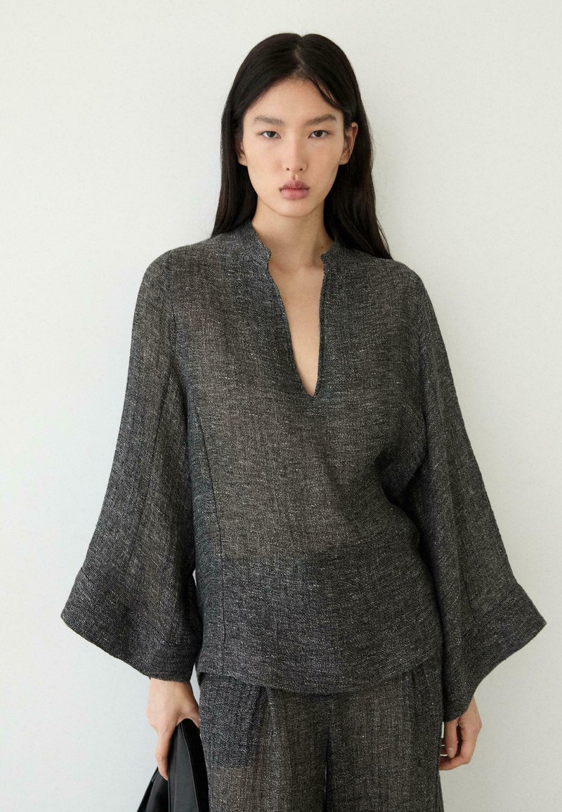 Kvinde med langt mørkt hår iført løs mørkegrå tekstureret top med brede ærmer og matchende bukser, der holder en sort genstand ved siden af sig.