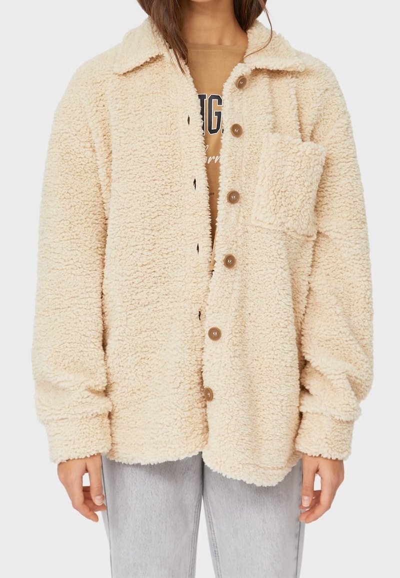 Veste en polaire sherpa beige avec boutons et poche poitrine portée sur un t-shirt graphique beige et un jean gris clair.