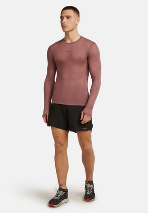 Mann steht da und trägt ein enganliegendes, langärmeliges, strukturiertes mauvefarbenes Oberteil, schwarze Shorts und schwarze Sneakers mit weißen und roten Sohlen vor einem weißen Hintergrund.