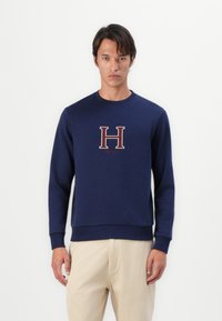 Felpa blu marina realizzata in tessuto morbido, con un ricamo maroon della lettera "H" sul petto, scollatura rotonda e polsini a costine.