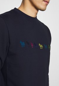 Granatowa bluza z haftowanymi zebrami w kolorach niebieskim, czarnym, różowym, żółtym i zielonym. Dekolt typu crew, tkanina wydaje się miękka i teksturowana.