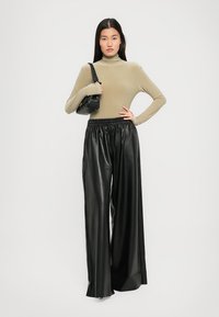 Donna che indossa un dolcevita lungo beige, pantaloni in pelle neri a vita alta e ampia, mentre tiene una borsa a tracolla nera, in piedi contro uno sfondo bianco.