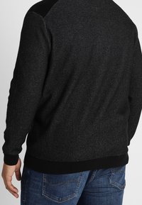 Maglione scuro con una texture e un design a maglia nera, caratterizzato da accenti sulle spalle in contrasto e polsini e orlo a coste. Indossato con jeans blu.