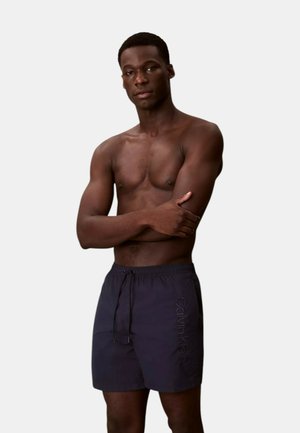 Uomo senza maglietta in piedi con le braccia incrociate, indossa pantaloncini da bagno Calvin Klein scuri su uno sfondo chiaro e uniforme.