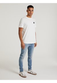 Weißes Baumwoll-T-Shirt mit kurzen Ärmeln, schwarzes Logopatch auf der Brust; kombiniert mit hellblauen Jeans und grauen Sneakers mit weißen Akzenten.