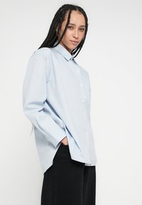 Gaiši zila garroku krekls ar pogām, brīvi pieguļošs, ar apkakli un sānu šķēlumiem, kopā ar melnām augsta jostasvietas biksēm.