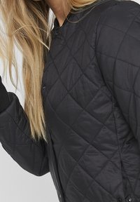 Giacca trapuntata nera, con un motivo a diamante, materiale testurizzato e polsini a maglia. Due bottoni a pressione visibili sul davanti.