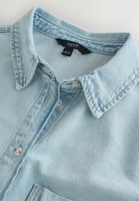 Col de chemise en denim bleu clair et patte de boutonnage, affichant l'étiquette de marque "NEXT" et l'étiquette "Made in Bangladesh" à l'intérieur du col.
