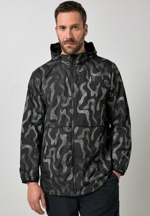 Mand iført sort hættetrøje med lys grå abstrakt prikket mønster, frontlynlås og lommer, stående mod ensartet baggrund.