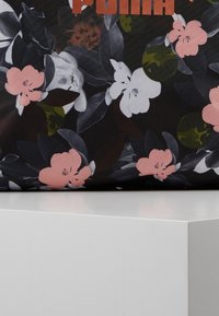 Kangas floral dengan bunga merah muda, putih, dan coklat serta daun gelap, menampilkan teks "PUMA" berwarna oranye yang terlihat sebagian di bagian atas.