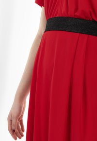 Rode jurk van gladde stof, met een zwarte glitter tailleband en zachte draping. Het kledingstuk is licht geplooid voor textuur.