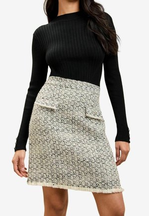 Kobieta ubrana w czarną, prążkowaną bluzkę z długim rękawem i wysoką, kremową spódnicę z tweedu z dwoma zapinanymi na guziki klapkowymi kieszeniami.