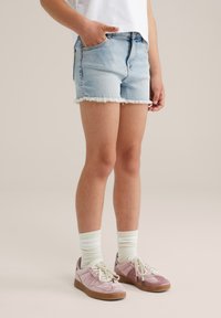 Hellblaue Denim-Shorts mit fransigem Saum, getragen mit cremefarbenen gestreiften Socken und rosa Wildledersneakern. Das Model hat eine Hand in der Tasche und präsentiert ein lässiges Outfit.