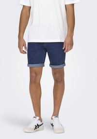 Shorts in denim blu scuro, con orli arrotolati, abbinati a una T-shirt bianca semplice e sneaker bianche con dettagli neri.