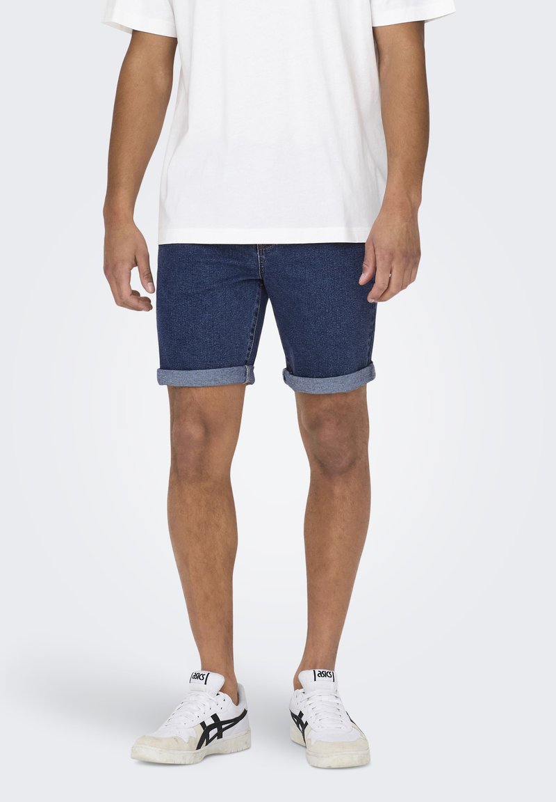 Shorts in denim blu scuro, con orli arrotolati, abbinati a una T-shirt bianca semplice e sneaker bianche con dettagli neri.