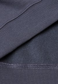 Tmavě šedá tkanina s hladkým pleteným texturováním a žebrovanými okraji, s jemným prošíváním podél švů.