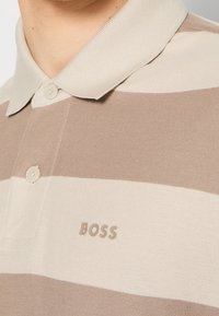 Polo à rayures beige et crème avec col boutonné et logo "BOSS" sur la poitrine, porté par une personne.