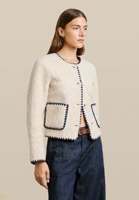 Valge tekstureeritud jakk tumesiniste äärtega, ümmarguse kaelusega, ees nupukesed ja kaks taskut. Kandmise all tume teksapükse.