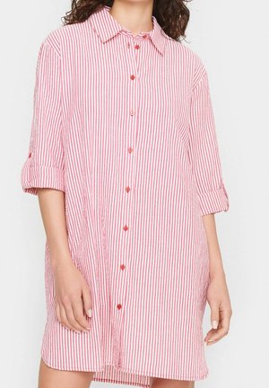 Vrouw draagt een losse, langarmige, roze-wit verticaal gestreepte overhemdjurk met opgerolde mouwen.